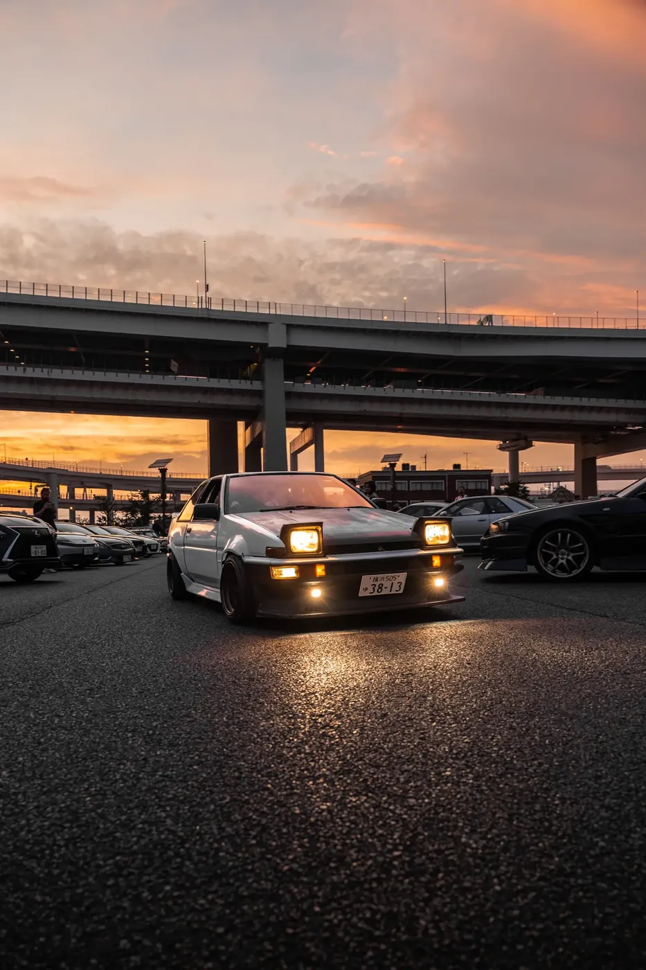 A white Toyota AE86 with its headlights on, parked beneath an elevated expressway at sunset.