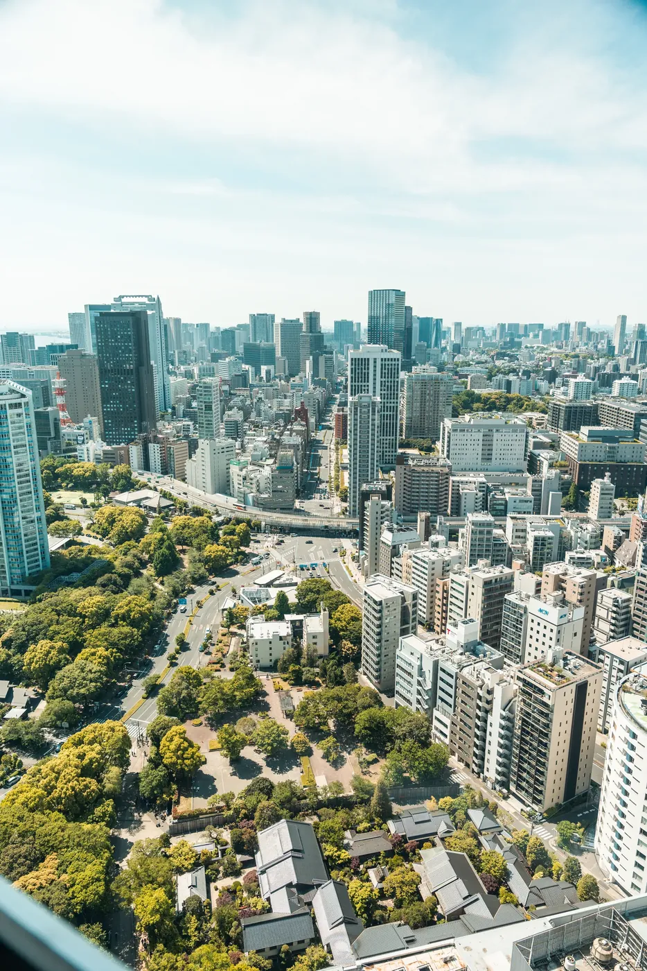 An aerial view over a dense city, tower blocks and a green park below a hazy skyline.