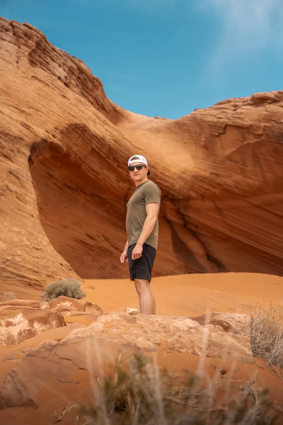A man in a backwards cap and sunglasses standing on red sandstone below a curved canyon wall under a clear blue sky.