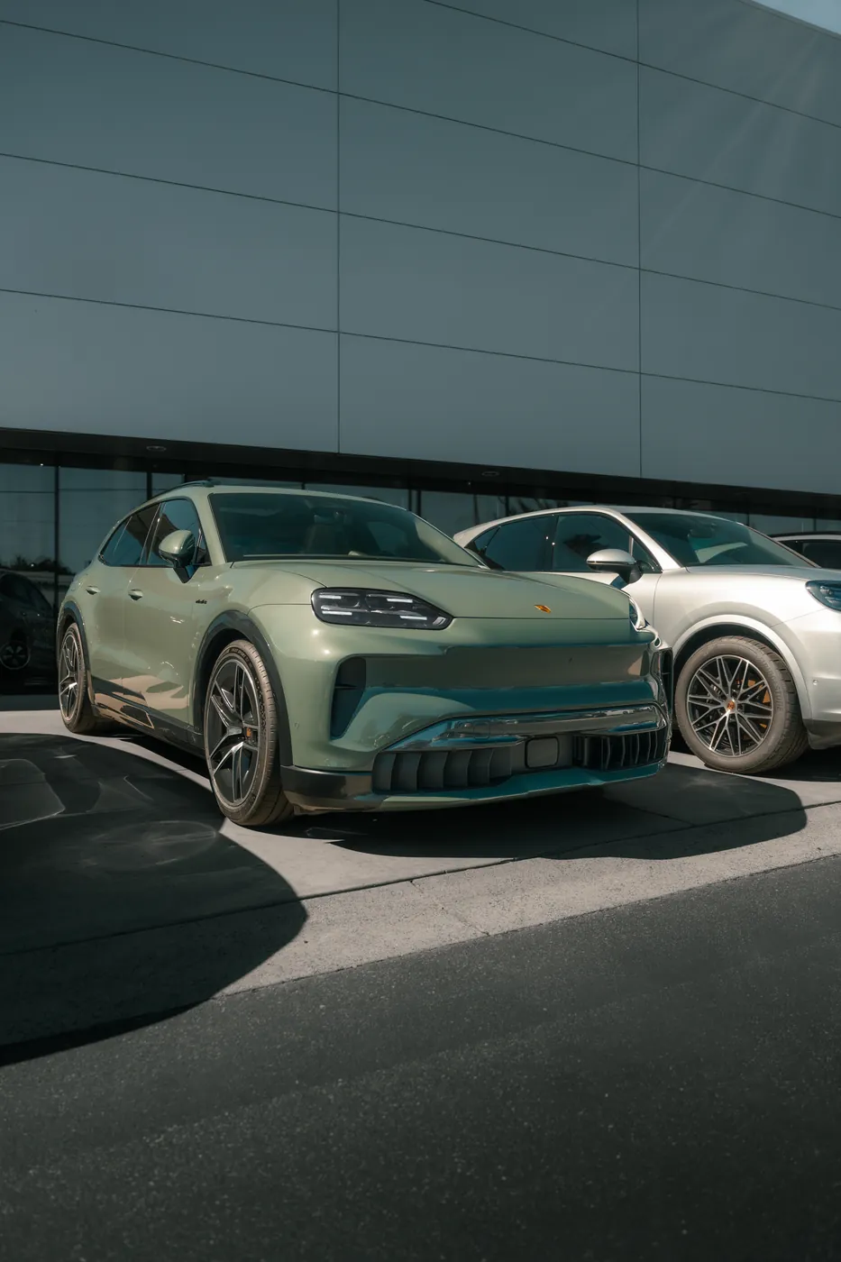 A sage green Porsche Macan parked outside a dealership in hard sunlight.