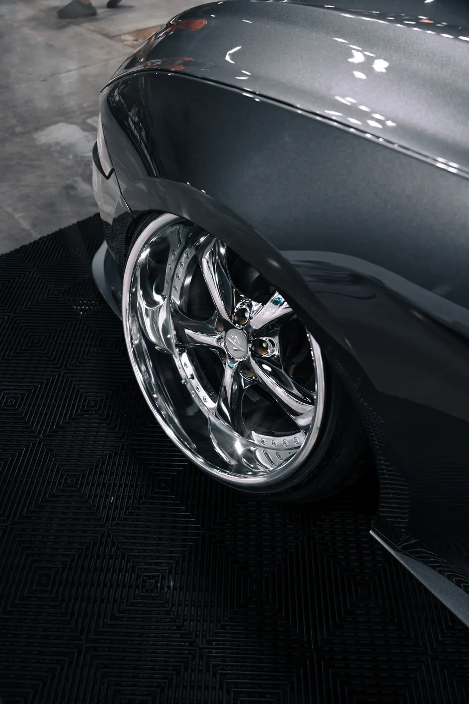 Close detail of a polished chrome wheel under a gray fender on a show floor.