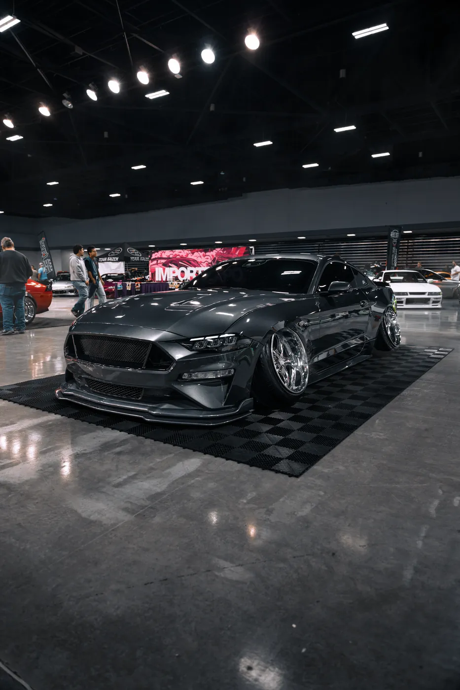 A gray Ford Mustang on chrome wheels, lowered onto a checkered display floor at an indoor car show.
