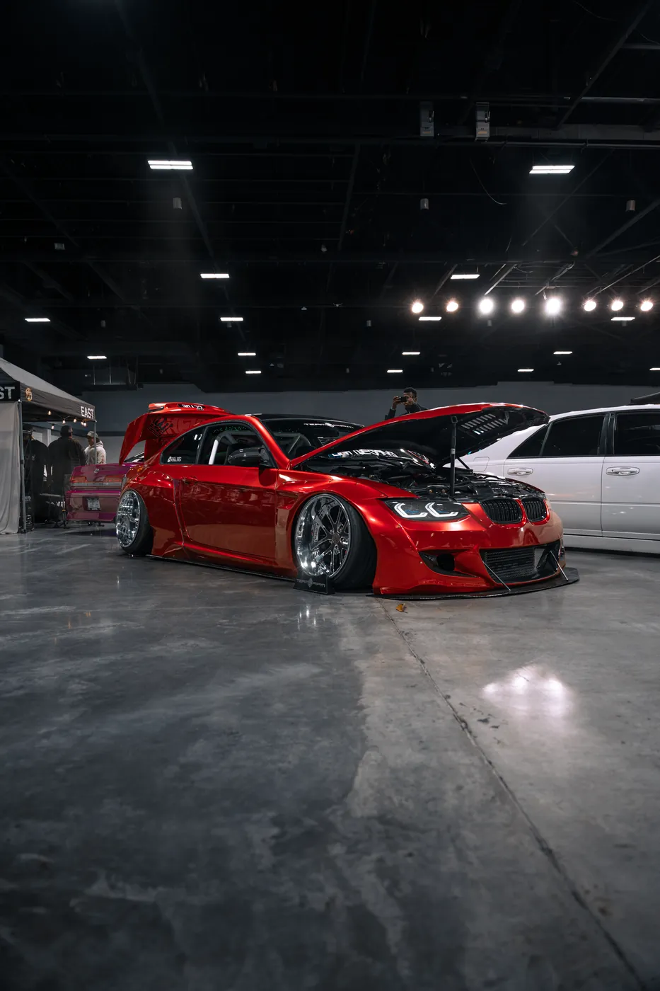 A candy red widebody BMW coupe with its hood and trunk open at an indoor car show.
