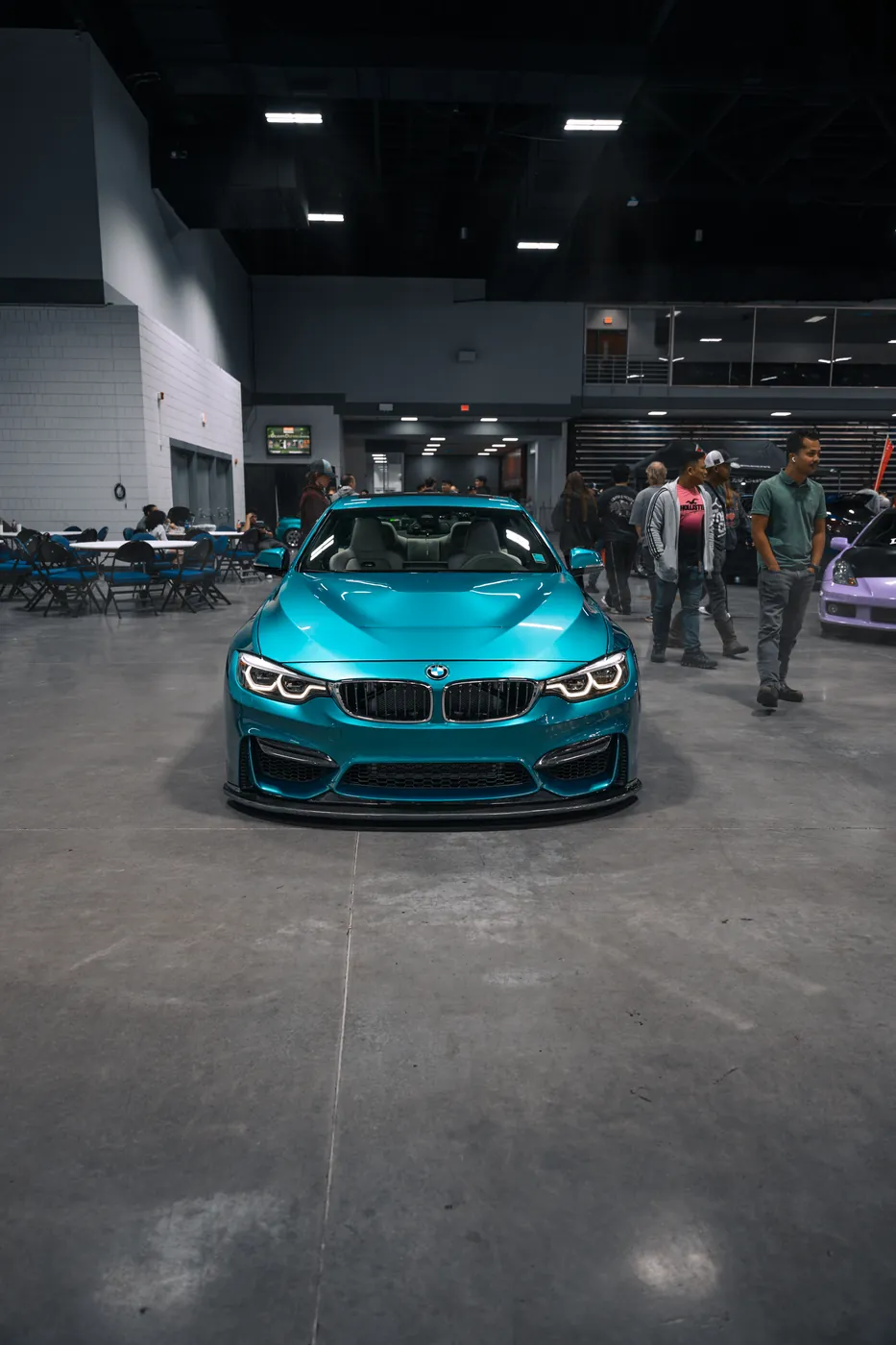 A teal BMW M4 shot head-on in a convention hall, show-goers behind it.
