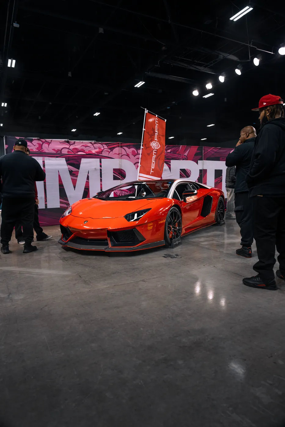 An orange Lamborghini Aventador in front of a pink mural at an import car show, spectators on both sides.