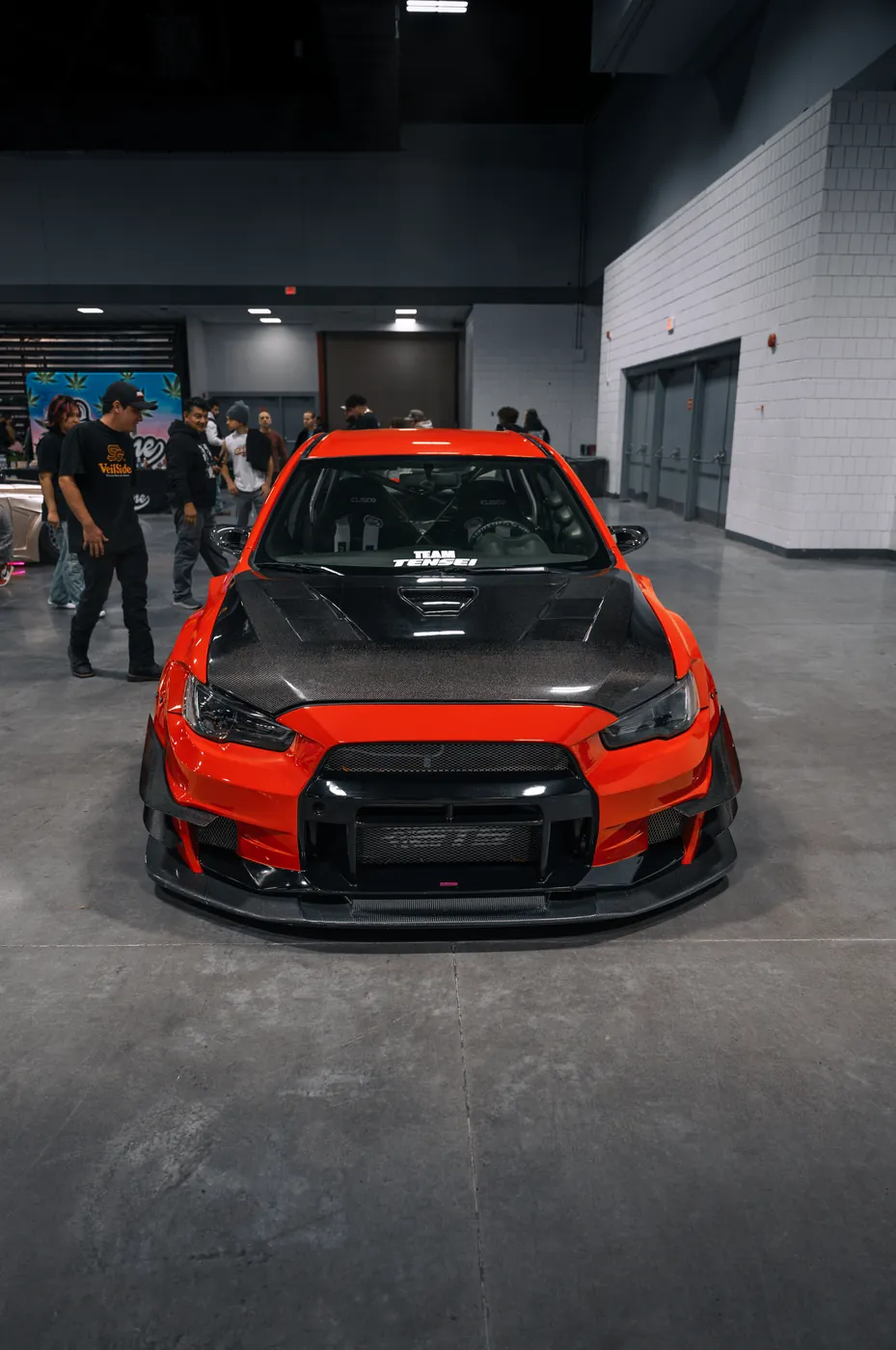 A red widebody Mitsubishi Evo with a carbon hood, shot head-on at an indoor car show.