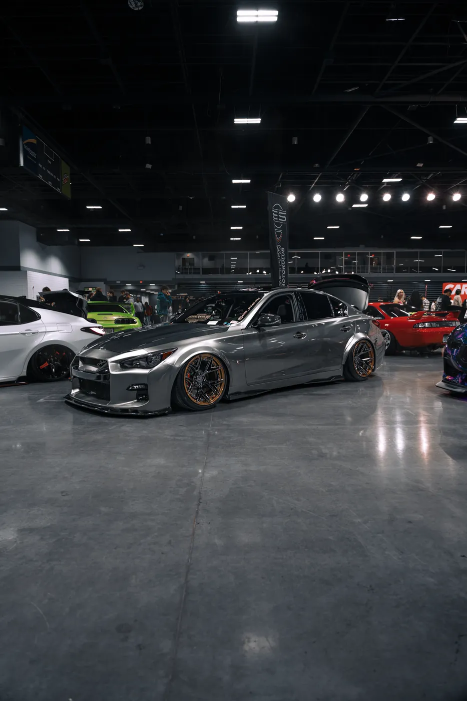 A silver Infiniti sedan on bronze-lipped wheels, lowered in a convention hall among other show cars.