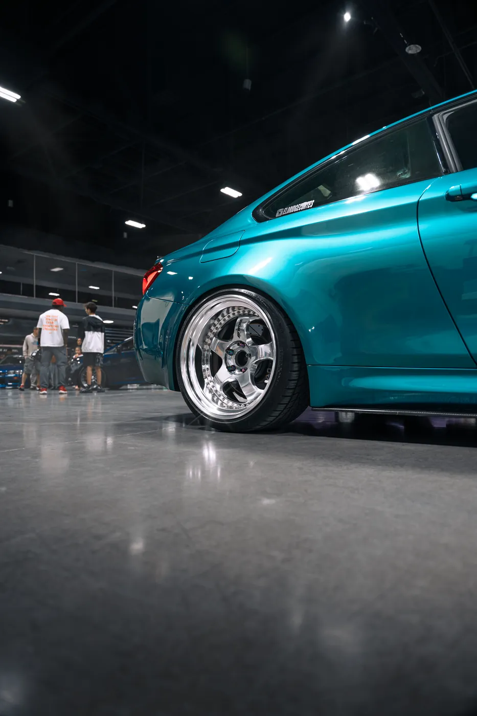 Low side view of a teal BMW M4 on polished five-spoke wheels at an indoor car show.