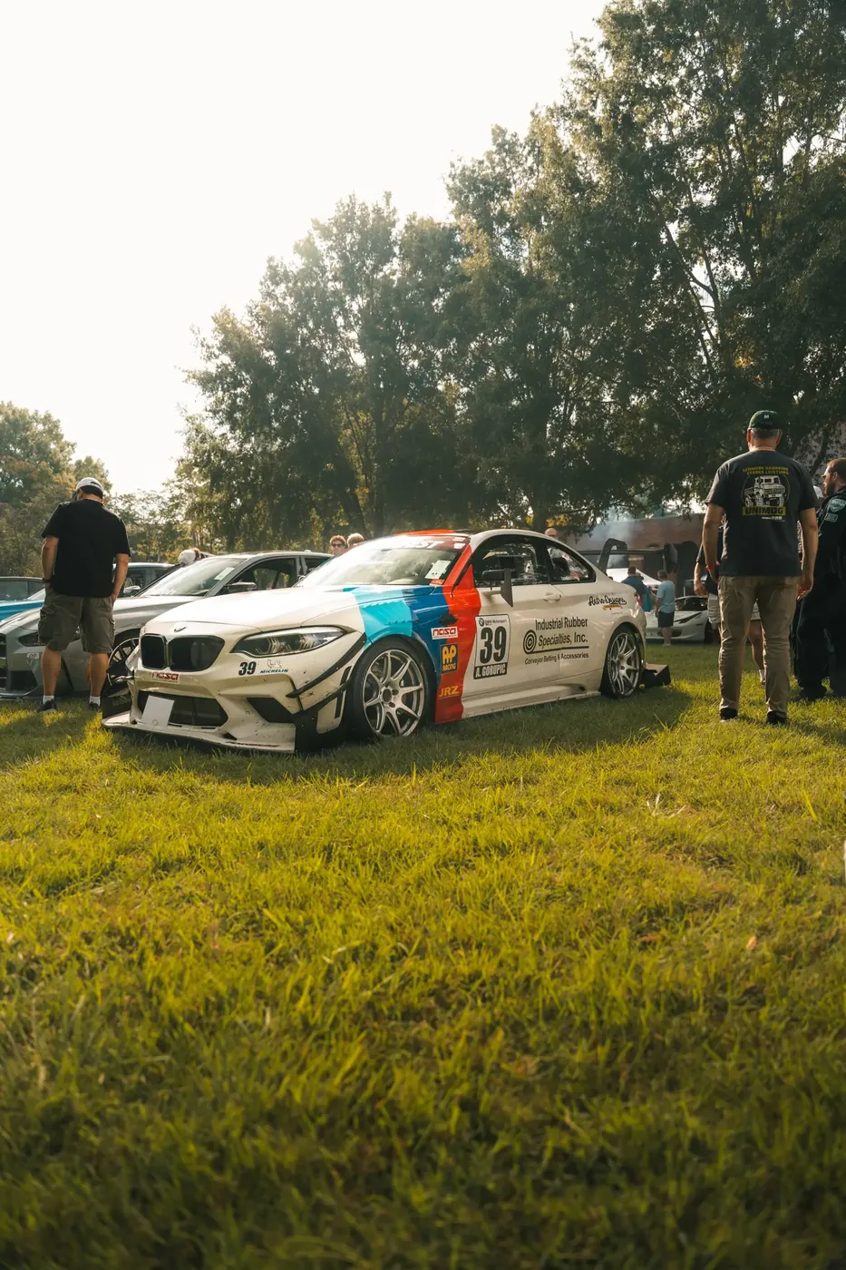 A white BMW M2 race car in motorsport stripes on a show lawn among spectators.