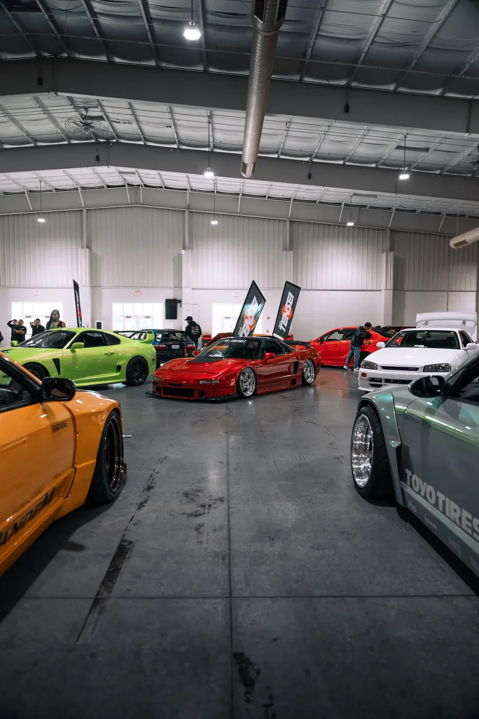 A red Acura NSX centered in an indoor show hall, framed by tuned Japanese cars.