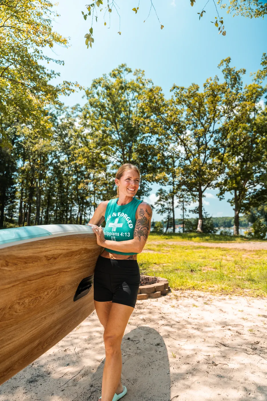 A woman carrying a wood-grain paddleboard on a sandy lakeshore, trees and water behind her.