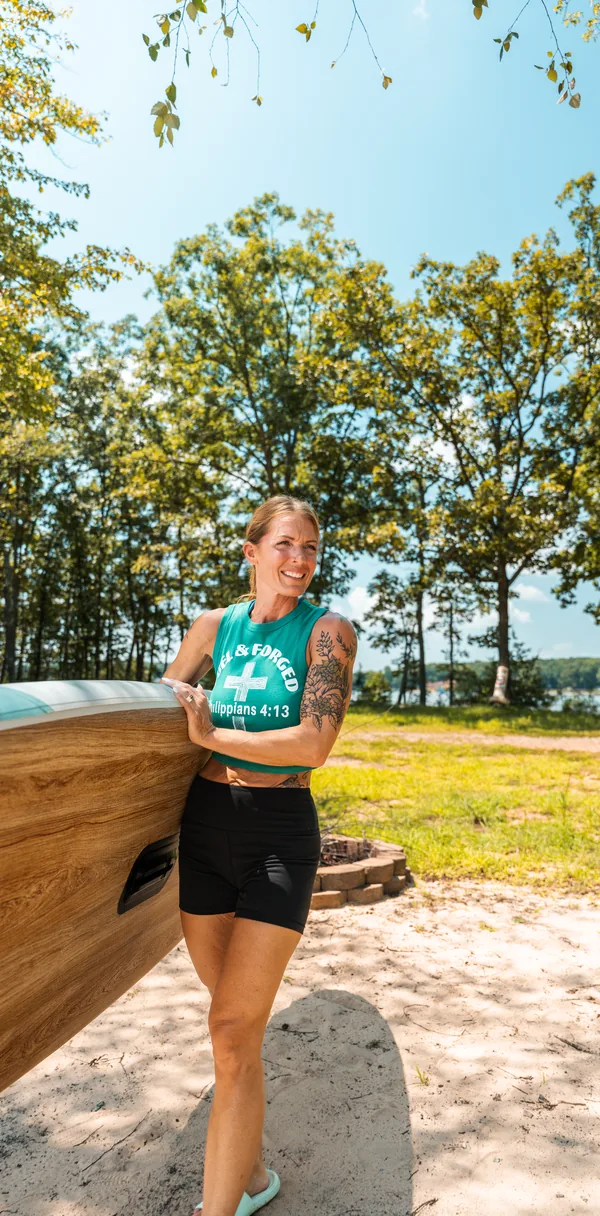 A woman carrying a wood-grain paddleboard on a sandy lakeshore, trees and water behind her.