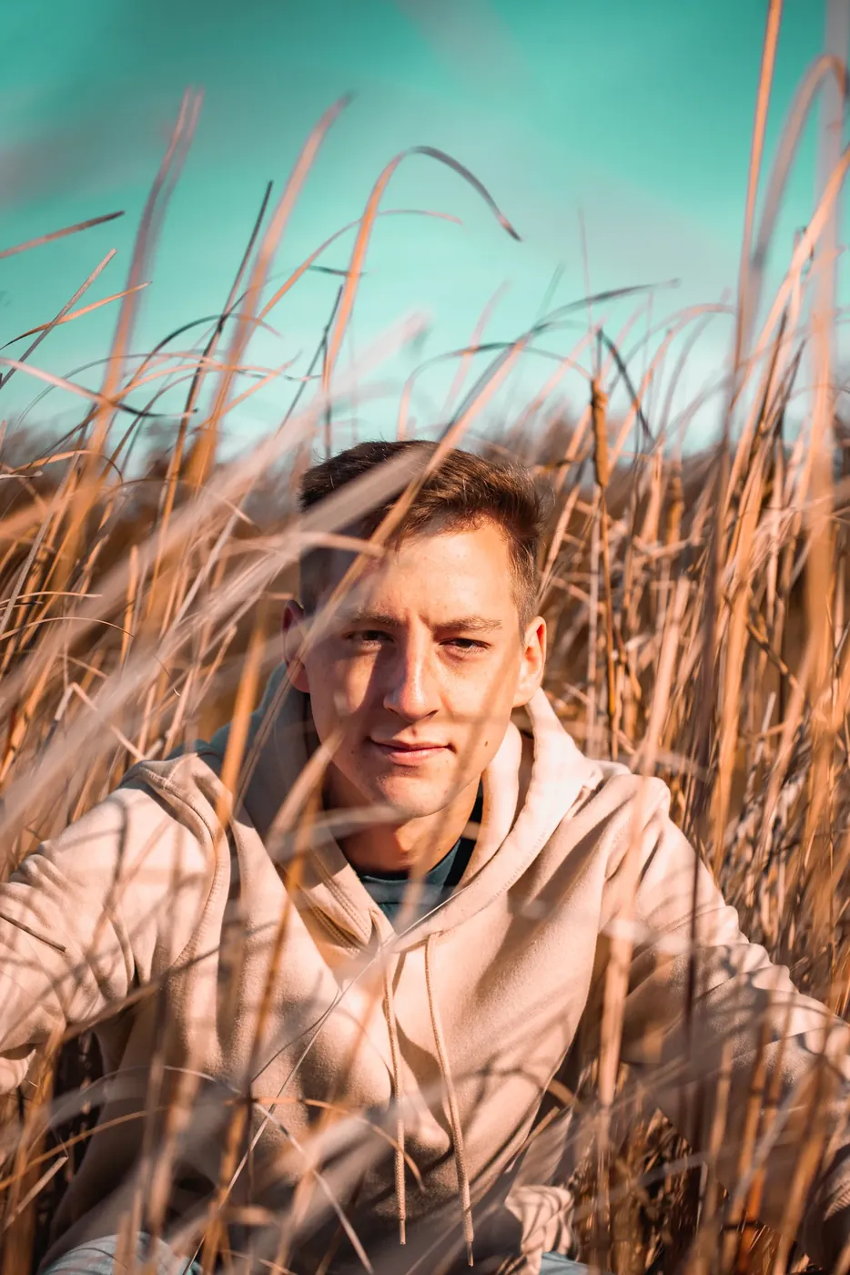 A man in a hooded sweatshirt photographed through tall dry grass.