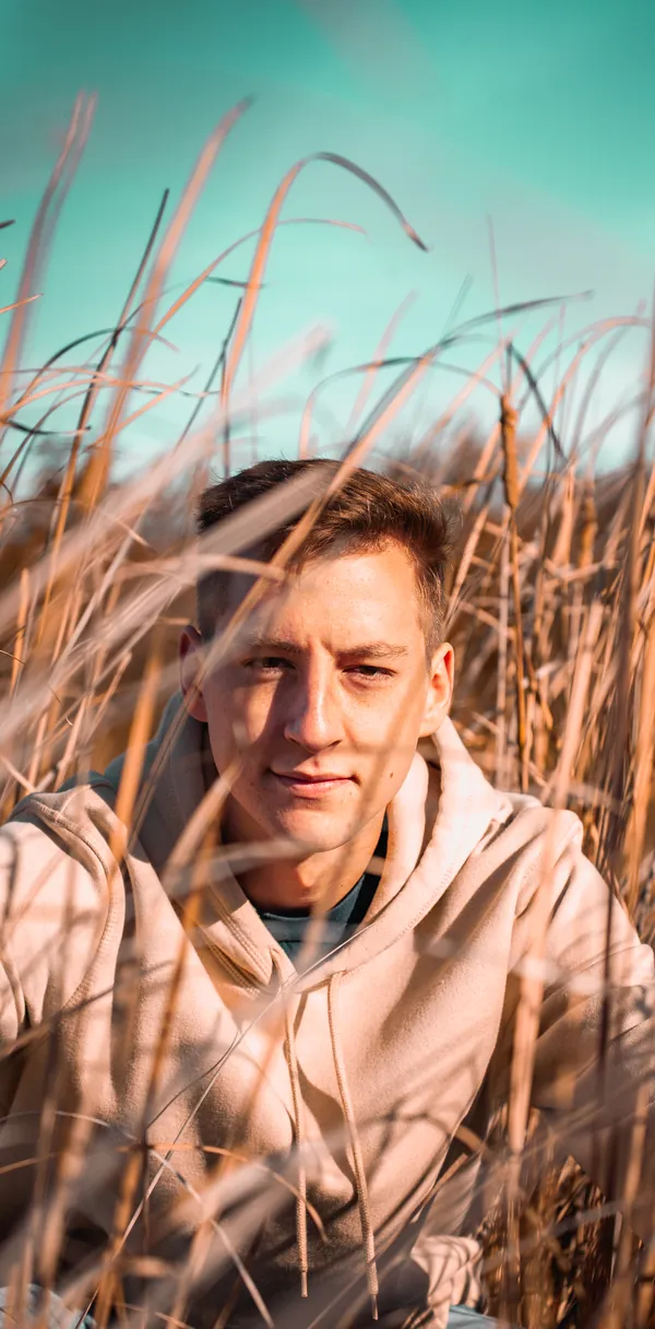 A man in a hooded sweatshirt photographed through tall dry grass.