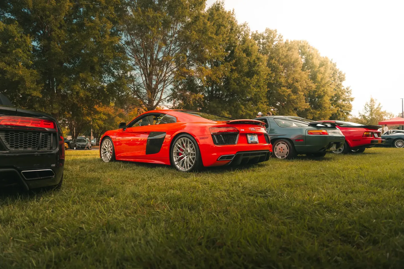 A red Audi R8 parked in a row with two Porsche 928s on a grassy show field.