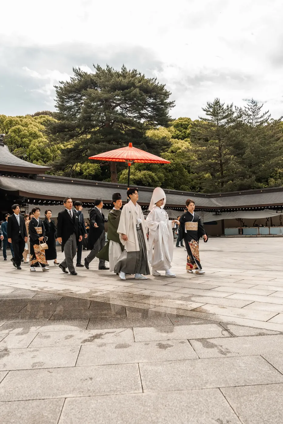 A traditional Shinto wedding procession crossing a shrine courtyard, the bride in white beneath a red parasol.