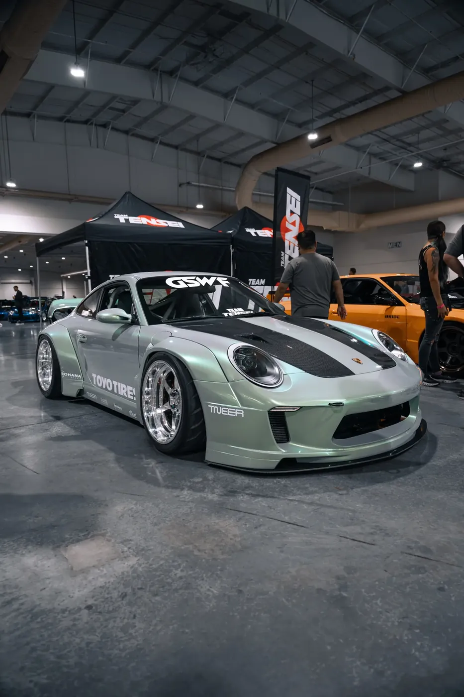 A mint-green widebody Porsche 911 on polished wheels at an indoor car show.
