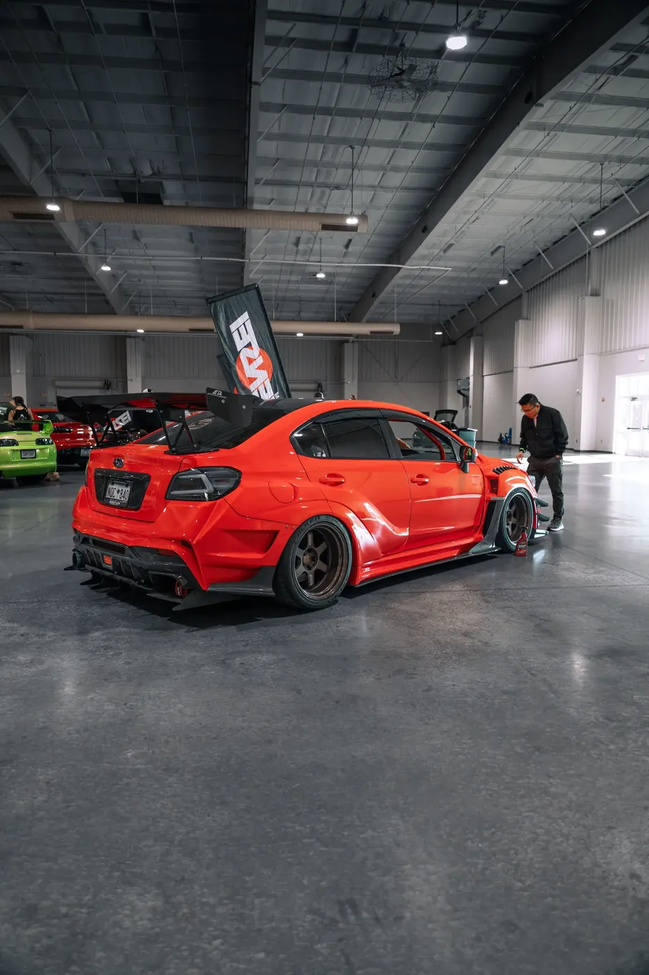 An orange widebody Subaru WRX with a tall rear wing, seen from behind in an indoor show hall.