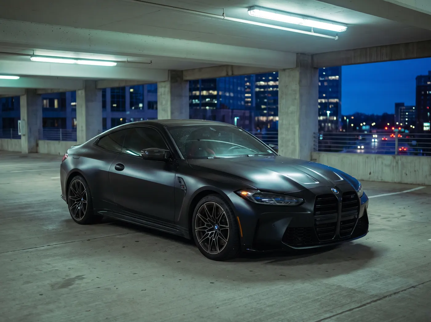 A BMW M4 wrapped in satin black vinyl, parked in a concrete parking structure at dusk.