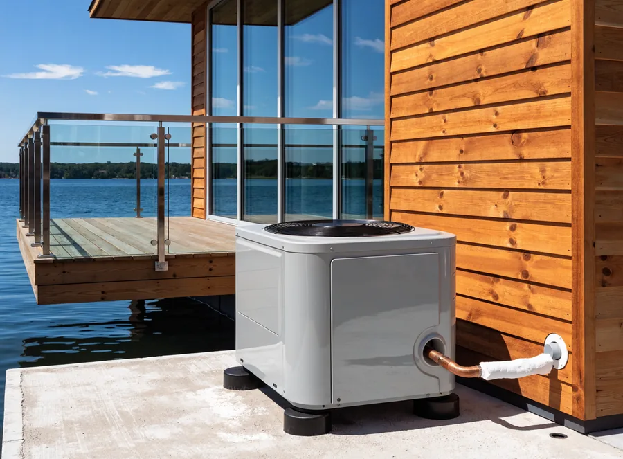 A newly installed condenser unit with tidy insulated line set beside a contemporary cedar-and-glass lakefront house, open lake water beyond.